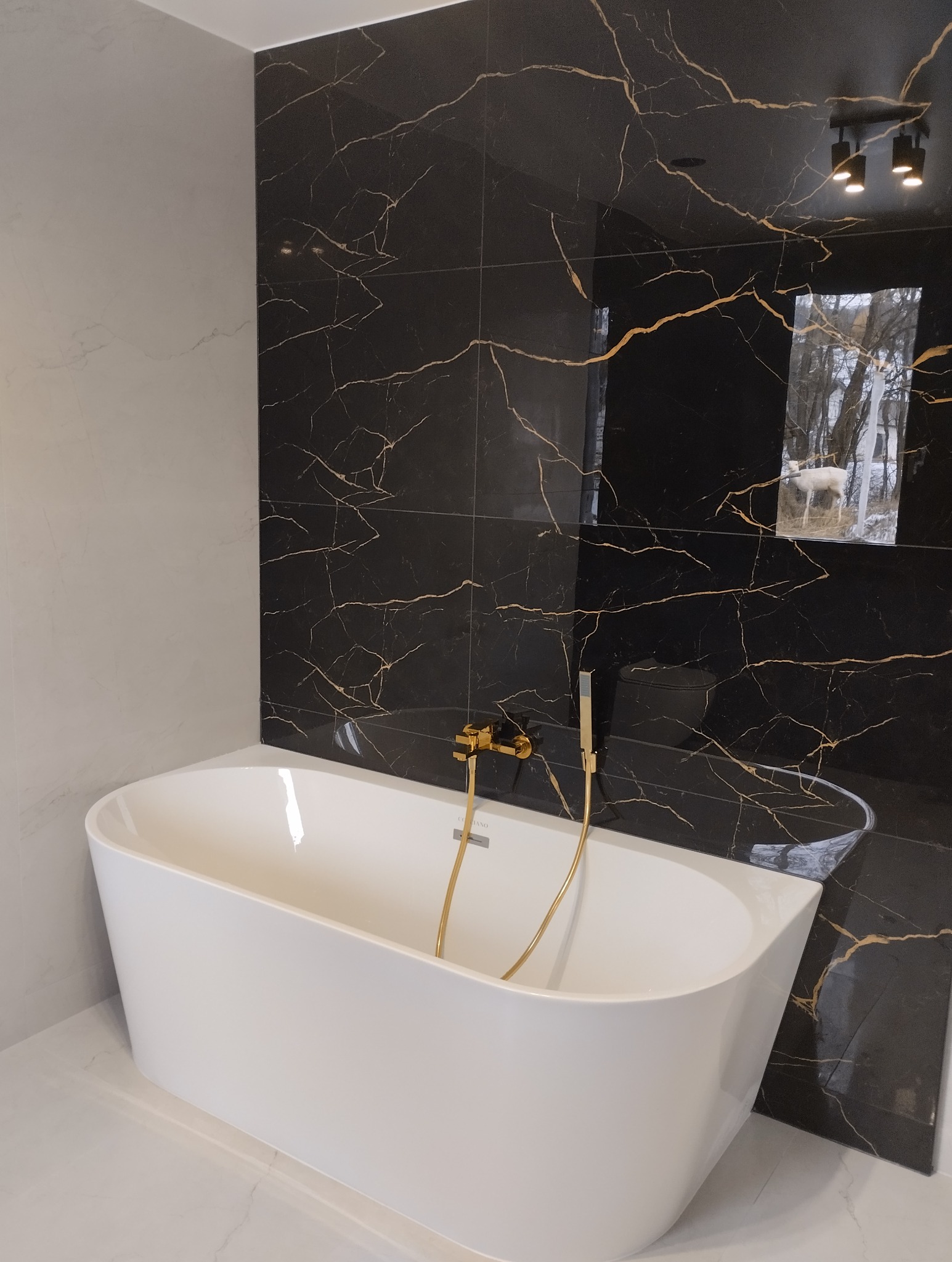 Nowoczesna łazienka z wolnostojącą wanną i czarnymi płytkami z złotymi żyłami. Bateria złota. W tle odbicie okna.