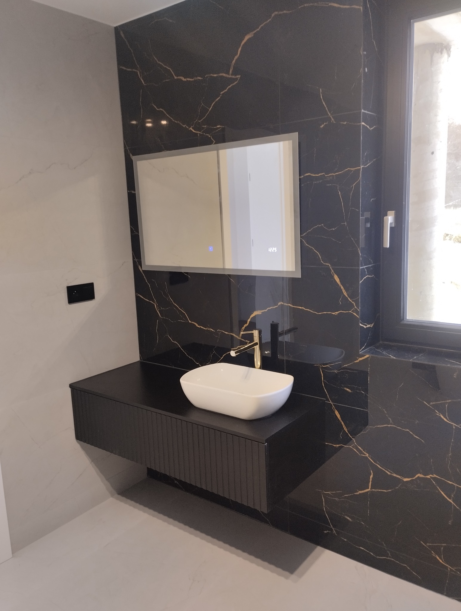 Elegancka łazienka z czarnymi płytkami imitującymi marmur, białą umywalką nablatową i podświetlanym lustrem. Minimalistyczna armatura w kolorze złotym.