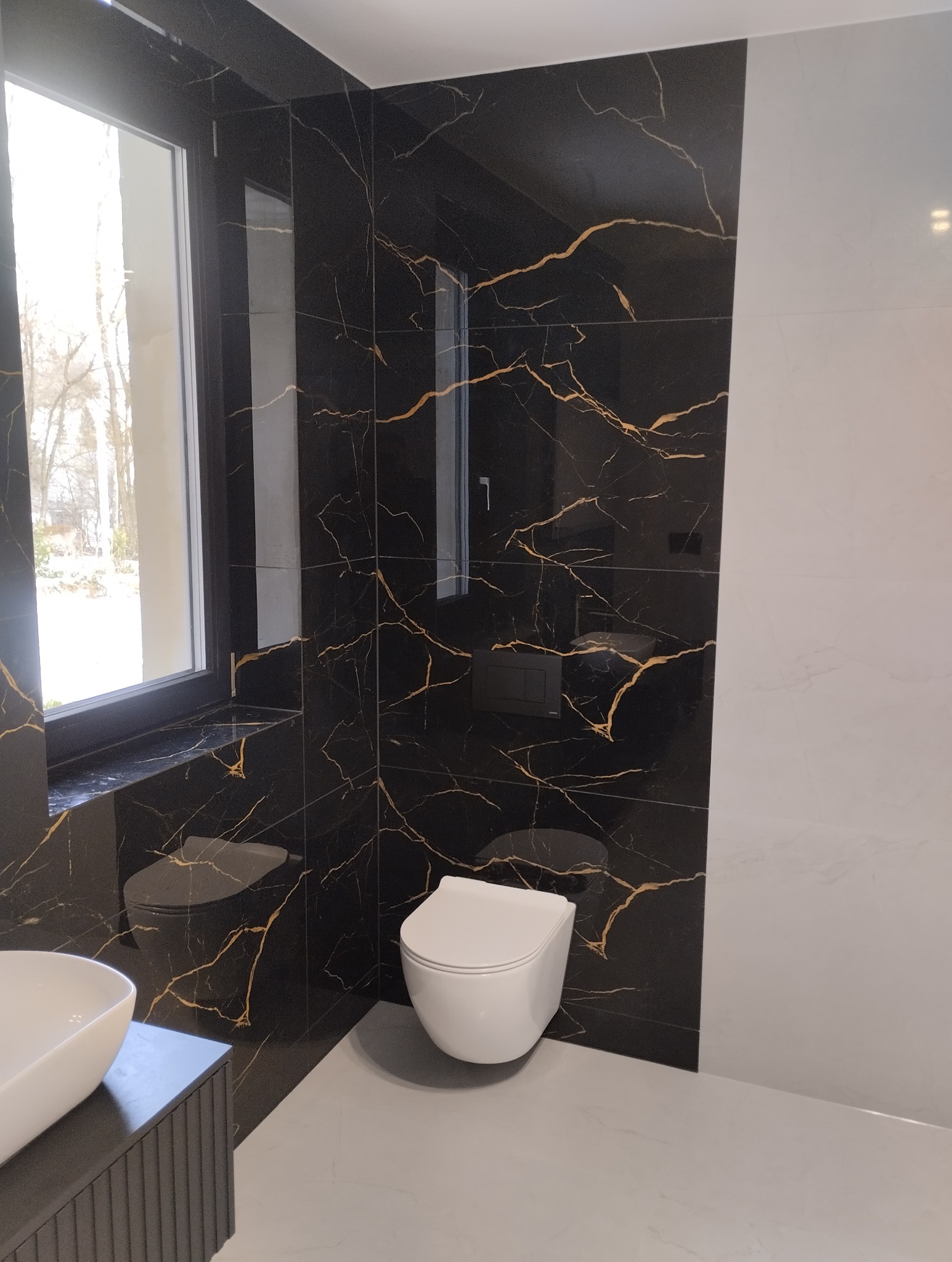 Elegancka łazienka z czarnymi płytkami imitującymi marmur ze złotymi żyłami, biała armatura i okno z widokiem na zewnątrz. Nowoczesny design.