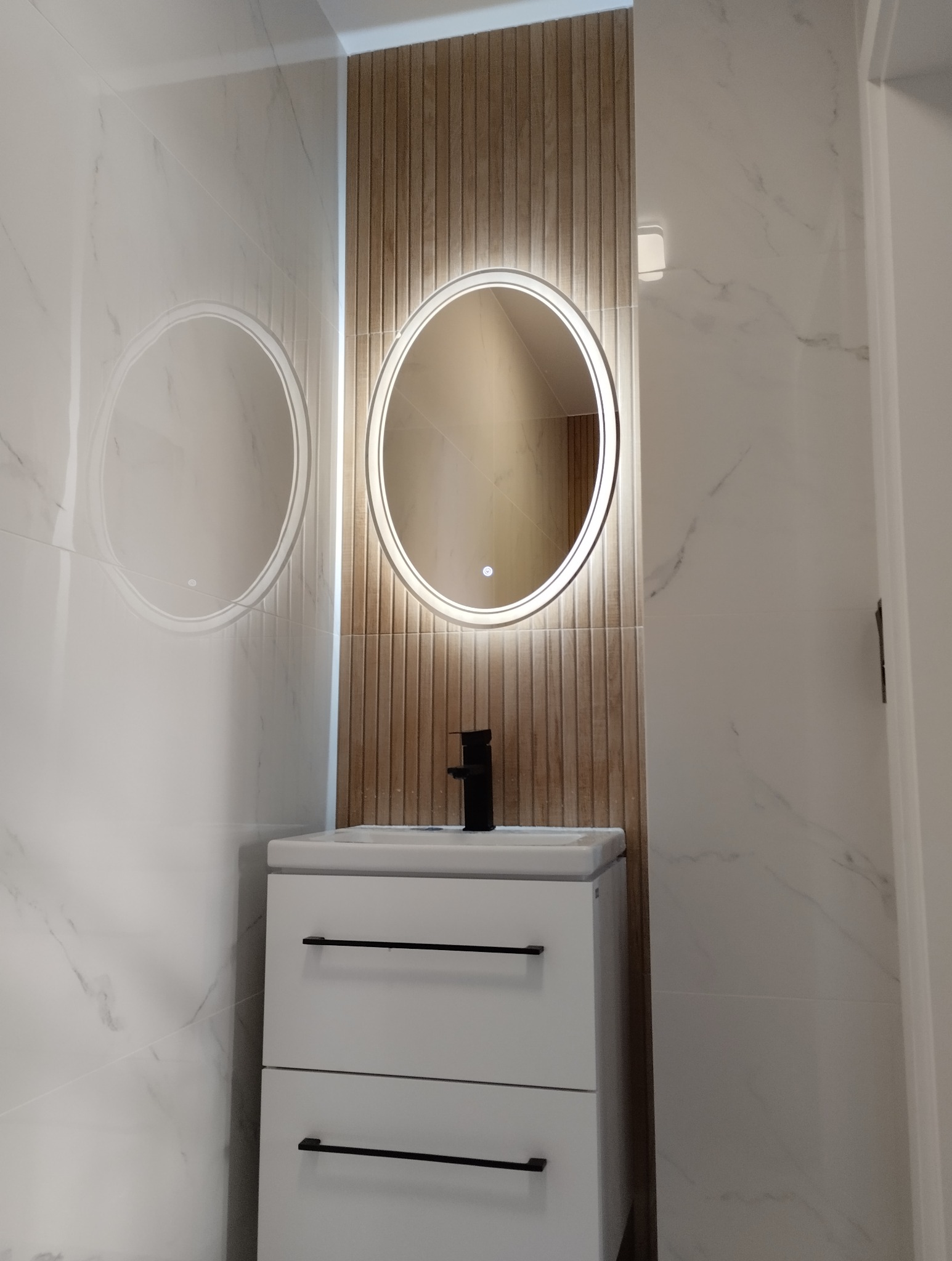 Nowoczesna łazienka: biała szafka z czarną armaturą pod owalnym lustrem z podświetleniem LED na tle drewnopodobnych paneli i marmurowych płytek.