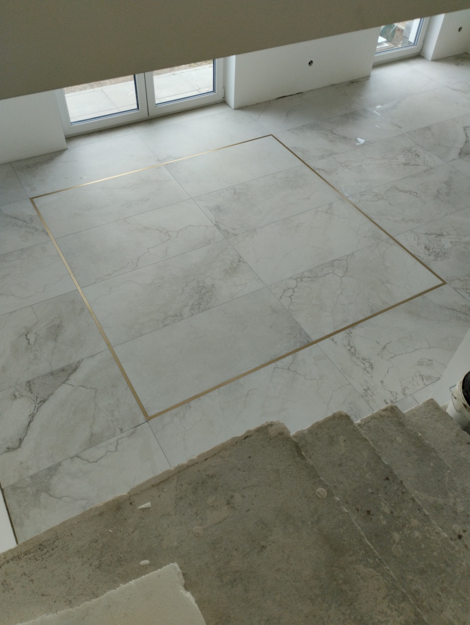 Jasne, marmurowe płytki podłogowe z dekoracyjną, złotą ramką, widok z góry schodów. Minimalistyczne wnętrze z oknami w tle.