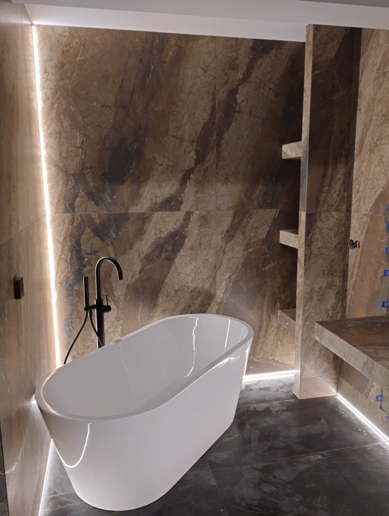 Nowoczesna łazienka z wolnostojącą białą wanną i marmurowymi płytkami. Podświetlenie LED podkreśla luksusowy charakter wnętrza. Czarna bateria wannowa.