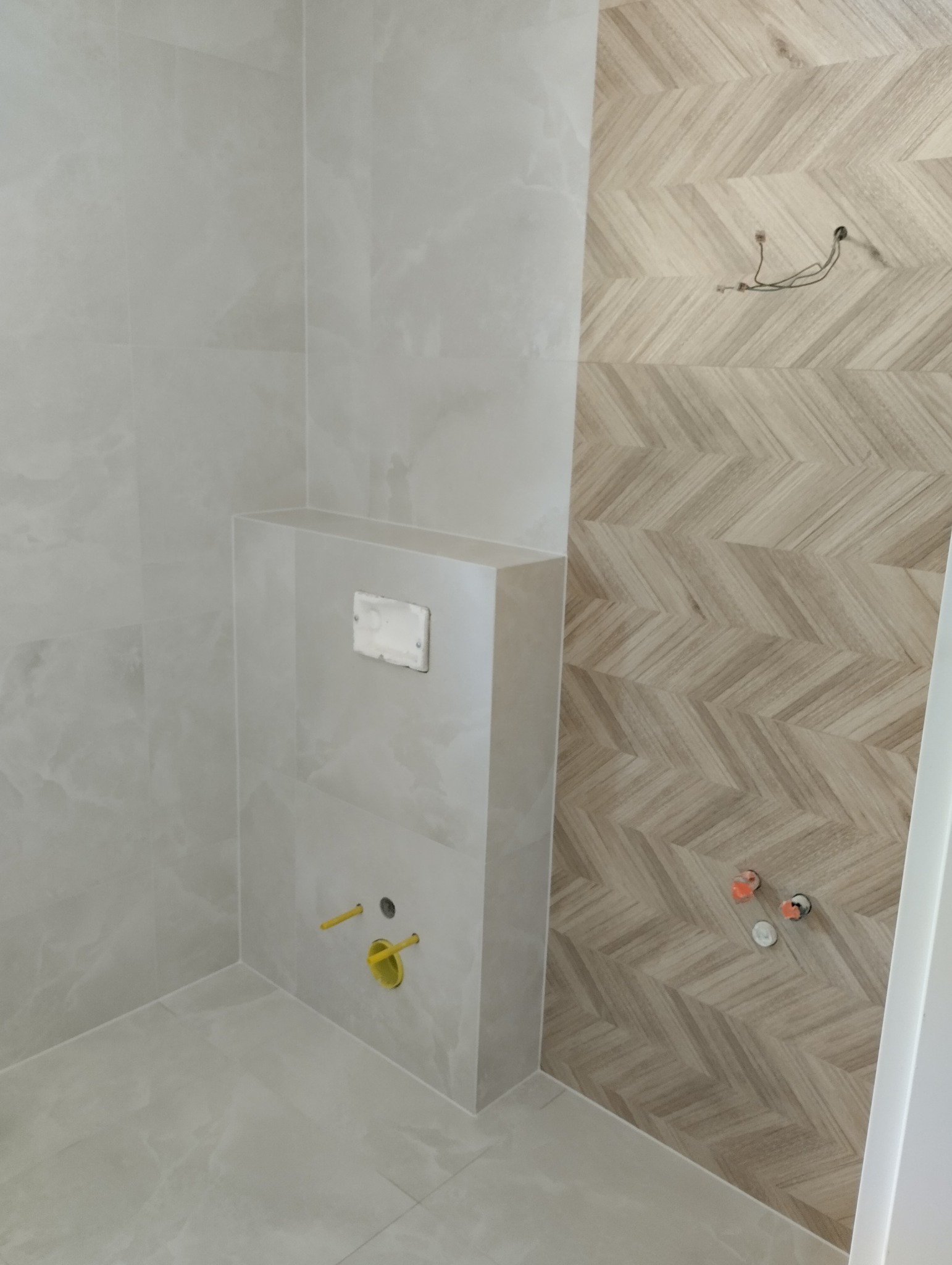 Wykończona łazienka z beżowymi płytkami ułożonymi w jodełkę na ścianie, widoczne przyłącza wodne i stelaż podtynkowy WC. Jasne, neutralne barwy.