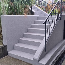 Oleg Bodnaruk BodChem - Schody zewnętrzne z szarego mikrocementu, z czarną metalową balustradą, otoczone zielenią. Widoczny fragment chodnika z kostki brukowej i drzwi rewizyjne.