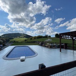 Oleg Bodnaruk BodChem - Jasnoszara posadzka żywiczna wokół basenu na zewnątrz, z widokiem na zielone wzgórza i błękitne niebo z chmurami. Osoba aplikuje materiał przy basenie.