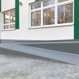 Oleg Bodnaruk BodChem - Zewnętrzny podjazd dla niepełnosprawnych z szarej wylewki, prowadzący do budynku z białą elewacją i zielonymi akcentami, kostka brukowa u podstawy.