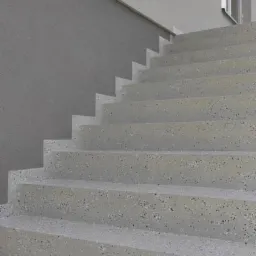 Schody zewnętrzne wykończone szarą, nakrapianą posadzką, prowadzące do budynku z szarą elewacją i oknem.