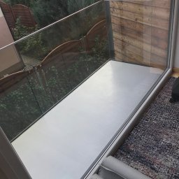 Oleg Bodnaruk BodChem - Balkon z szarą, gładką powierzchnią podłogi, otoczony szklaną balustradą. Widoczne rolki zabezpieczające podłogę i dywan w tle.