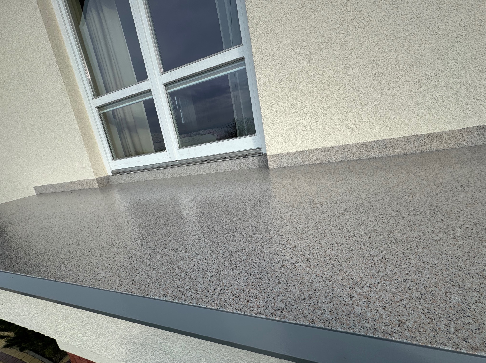Szara posadzka żywiczna z drobnym wzorem, wykonana na balkonie przy oknie. Widoczna krawędź balkonu i fragment elewacji budynku. Gładka powierzchnia, nowoczesny wygląd.