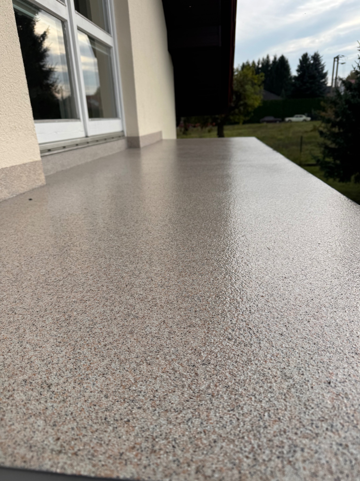 Szara posadzka żywiczna na balkonie z widokiem na zieleń. Gładka powierzchnia z delikatnym wzorem, okno balkonowe w tle. Nowoczesne wykończenie.