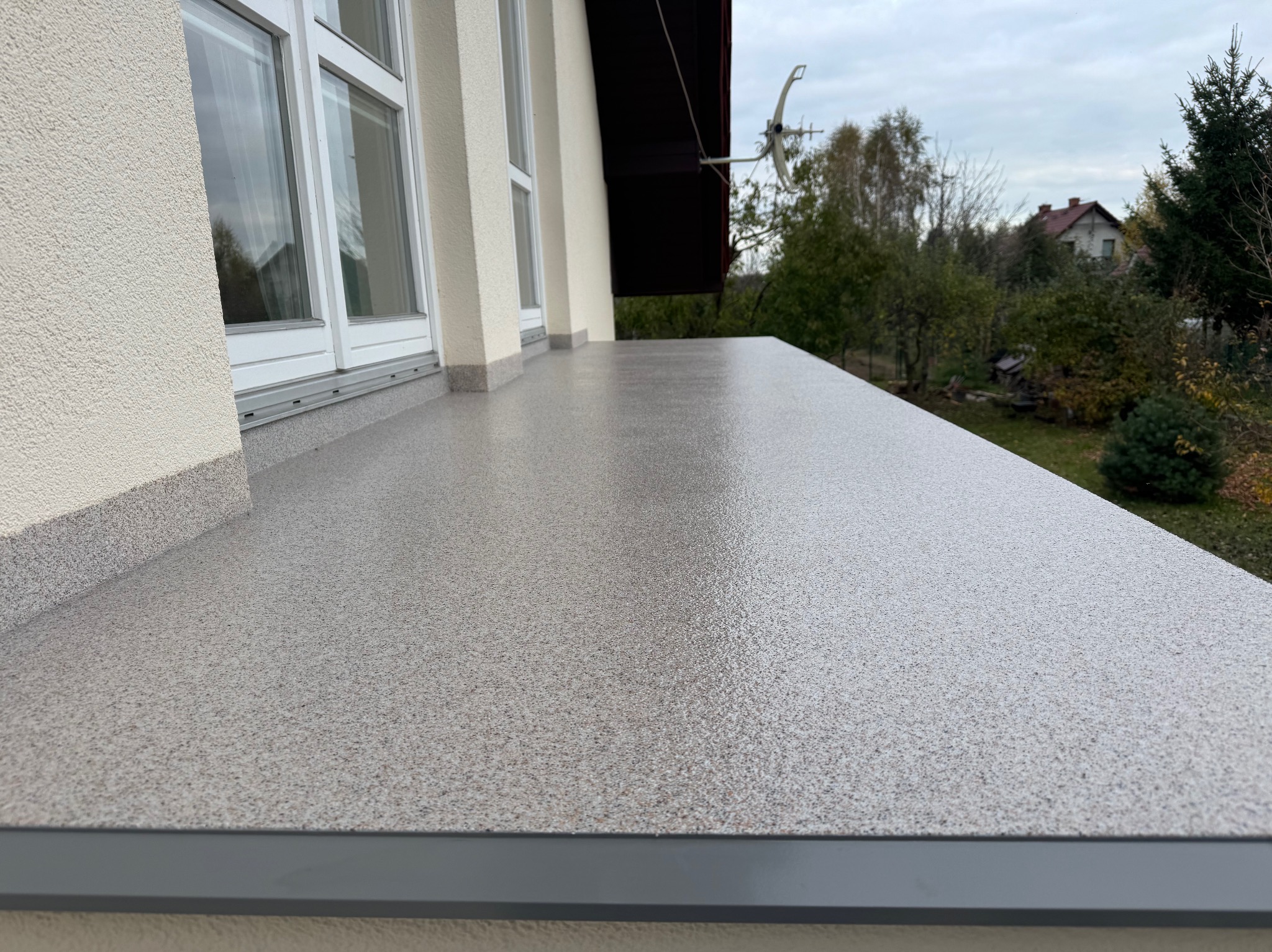 Balkon z szarą, gładką posadzką żywiczną w Krakowie. Widoczne okna i fragment elewacji budynku. W tle zieleń drzew i zabudowa jednorodzinna.