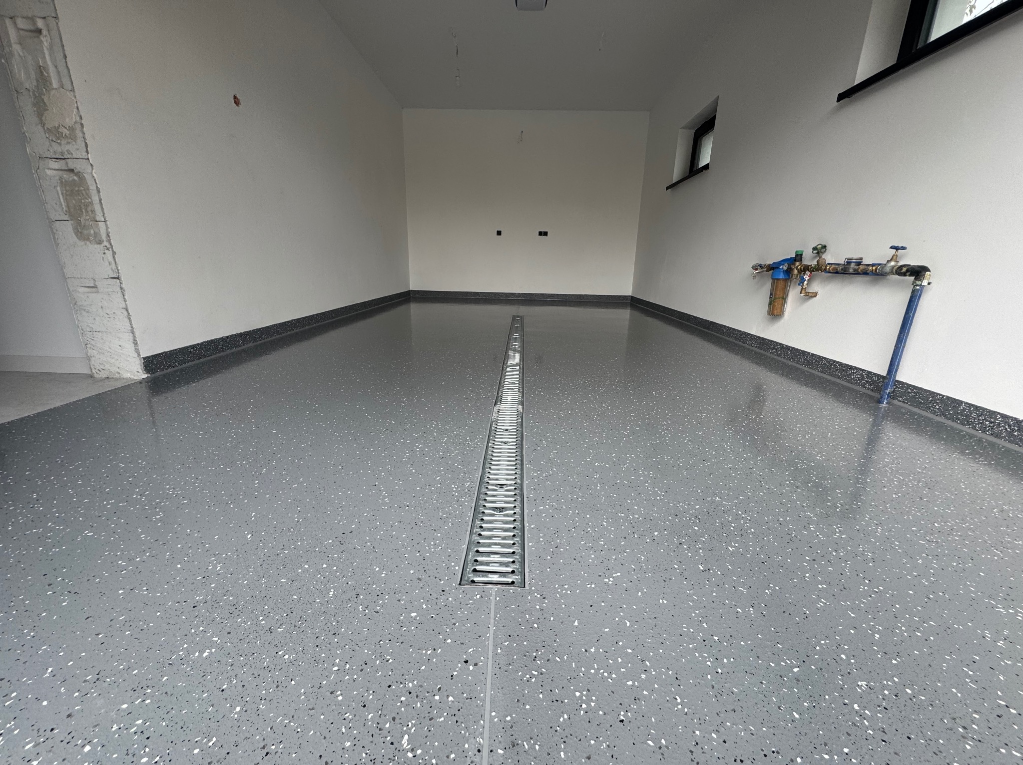 Szara posadzka żywiczna z odpływem liniowym w garażu. Widoczne białe ściany i instalacja wodna. Nowoczesne wykończenie podłogi z efektem lastryko.