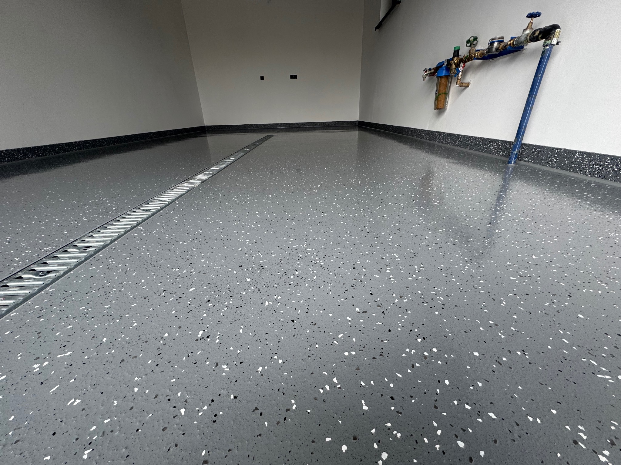 Szara posadzka żywiczna z odpływem liniowym w garażu, widoczne instalacje na ścianie. Błyszcząca powierzchnia z drobnymi, białymi i czarnymi elementami.