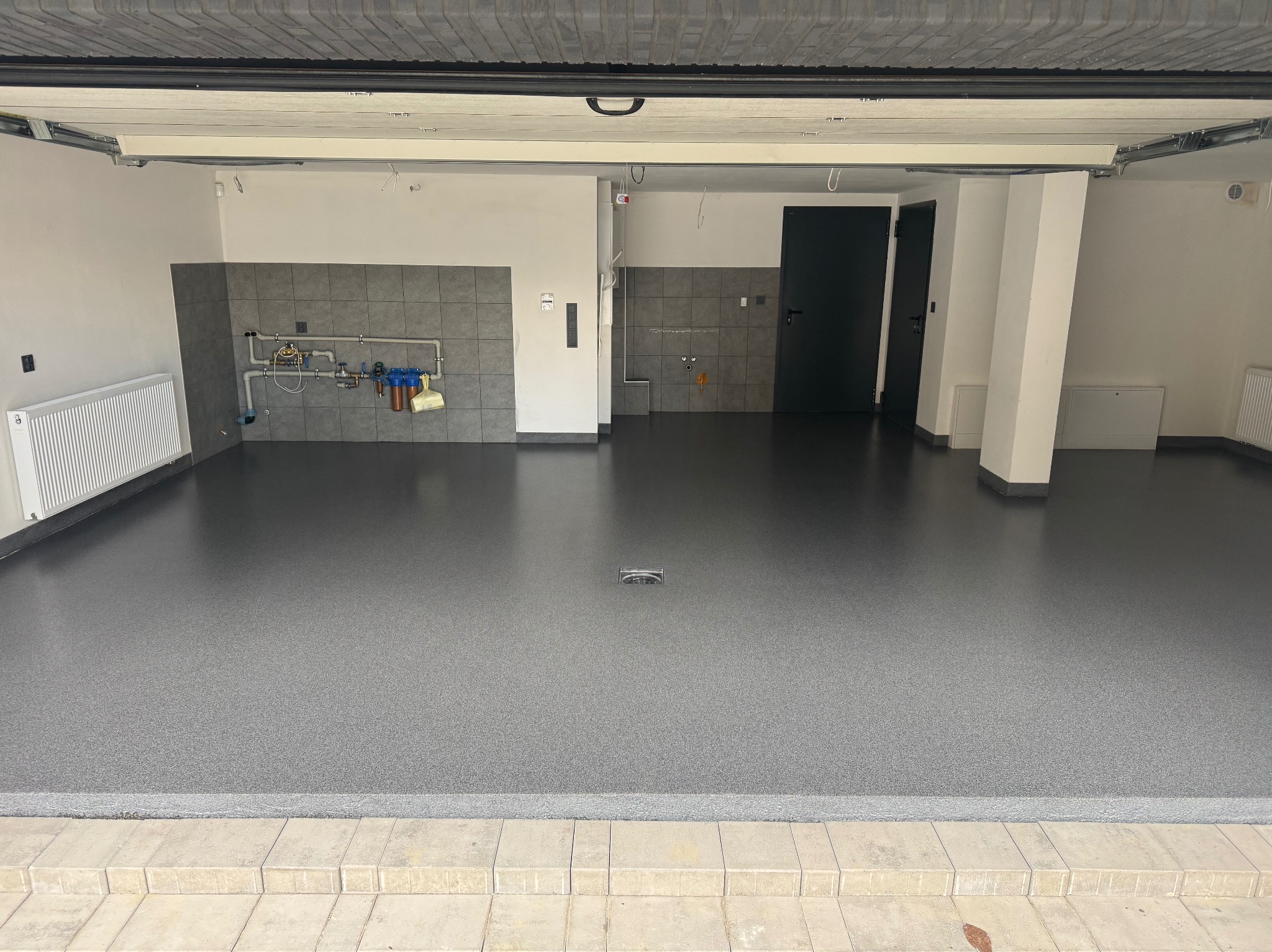 Gładka, szara posadzka żywiczna w garażu z szarą glazurą na ścianach. Widoczny odpływ liniowy i instalacja wodna. Ujęcie z progu garażu.