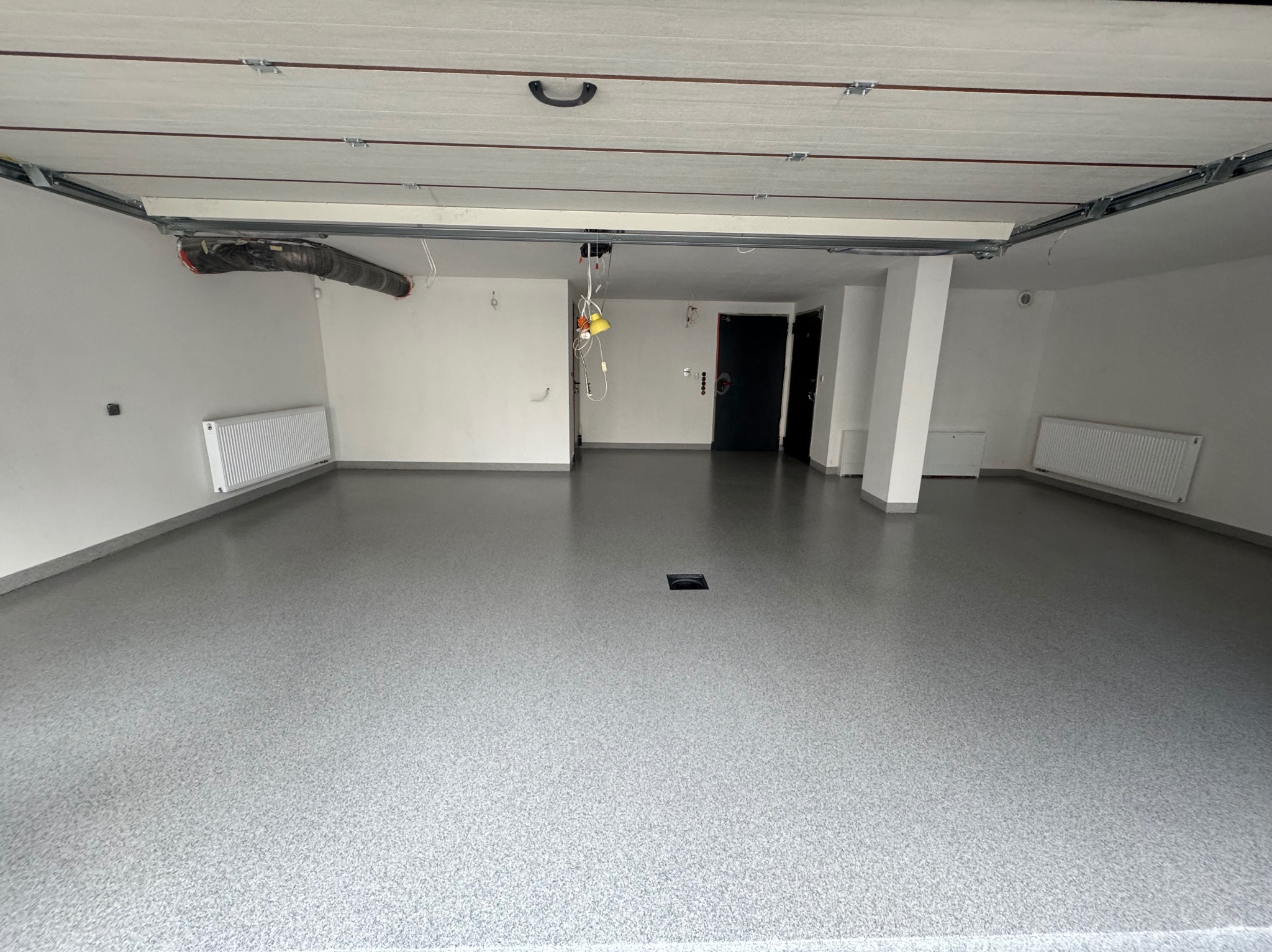 Jasnoszara posadzka żywiczna w garażu z grzejnikami i instalacją pod sufitem. Widok na gładką, jednolitą powierzchnię z delikatnym wzorem.