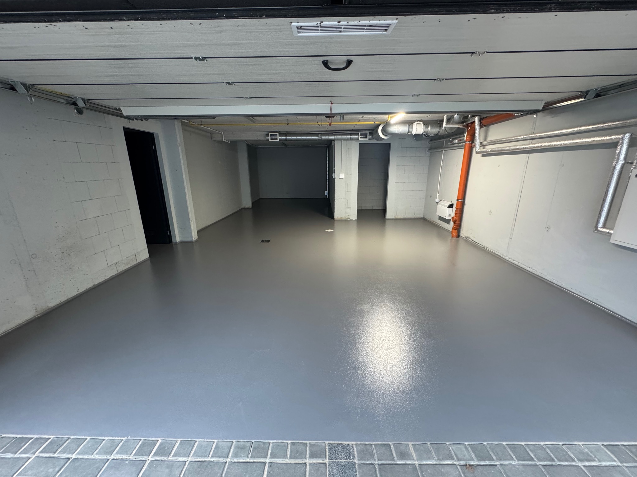 Gładka, szara posadzka żywiczna w garażu z betonowymi ścianami i instalacjami pod sufitem. Widok na wnętrze garażu z otwartymi drzwiami.