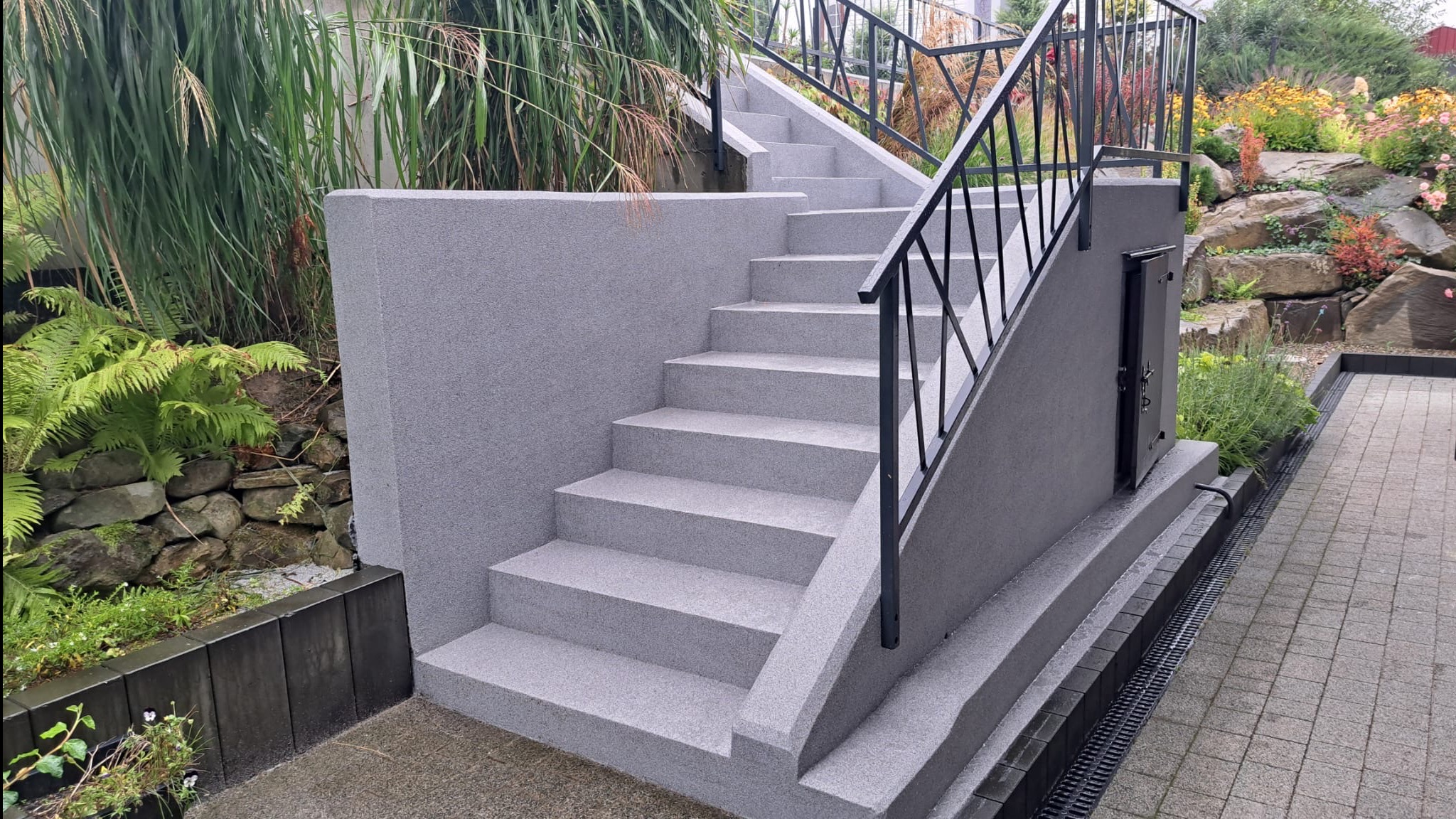 Schody zewnętrzne z szarego mikrocementu, z czarną metalową balustradą, otoczone zielenią. Widoczny fragment chodnika z kostki brukowej i drzwi rewizyjne.
