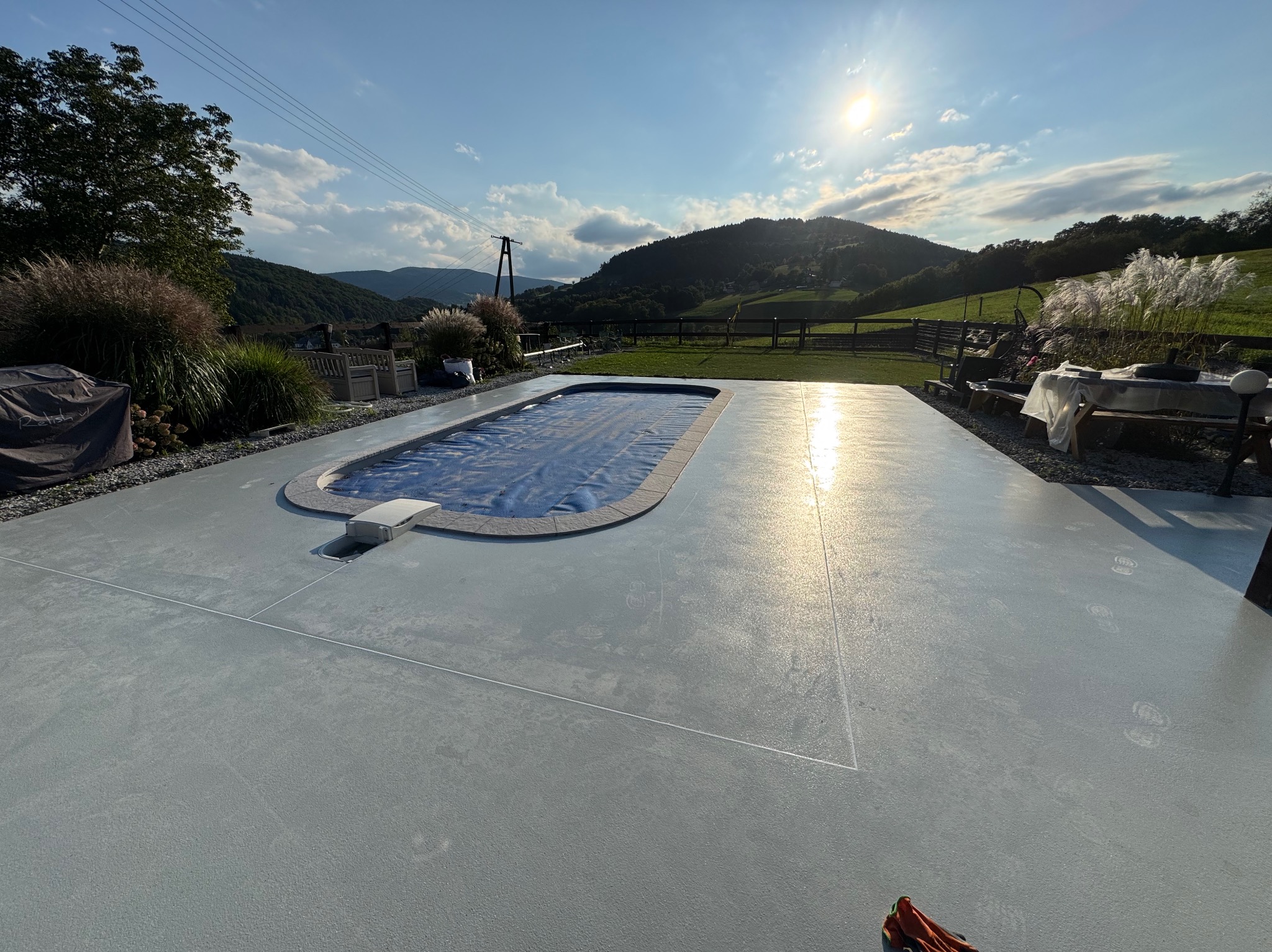 Szara posadzka wokół basenu z widokiem na góry w słoneczny dzień. Nowoczesna powierzchnia z delikatnym połyskiem i widocznymi śladami stóp.
