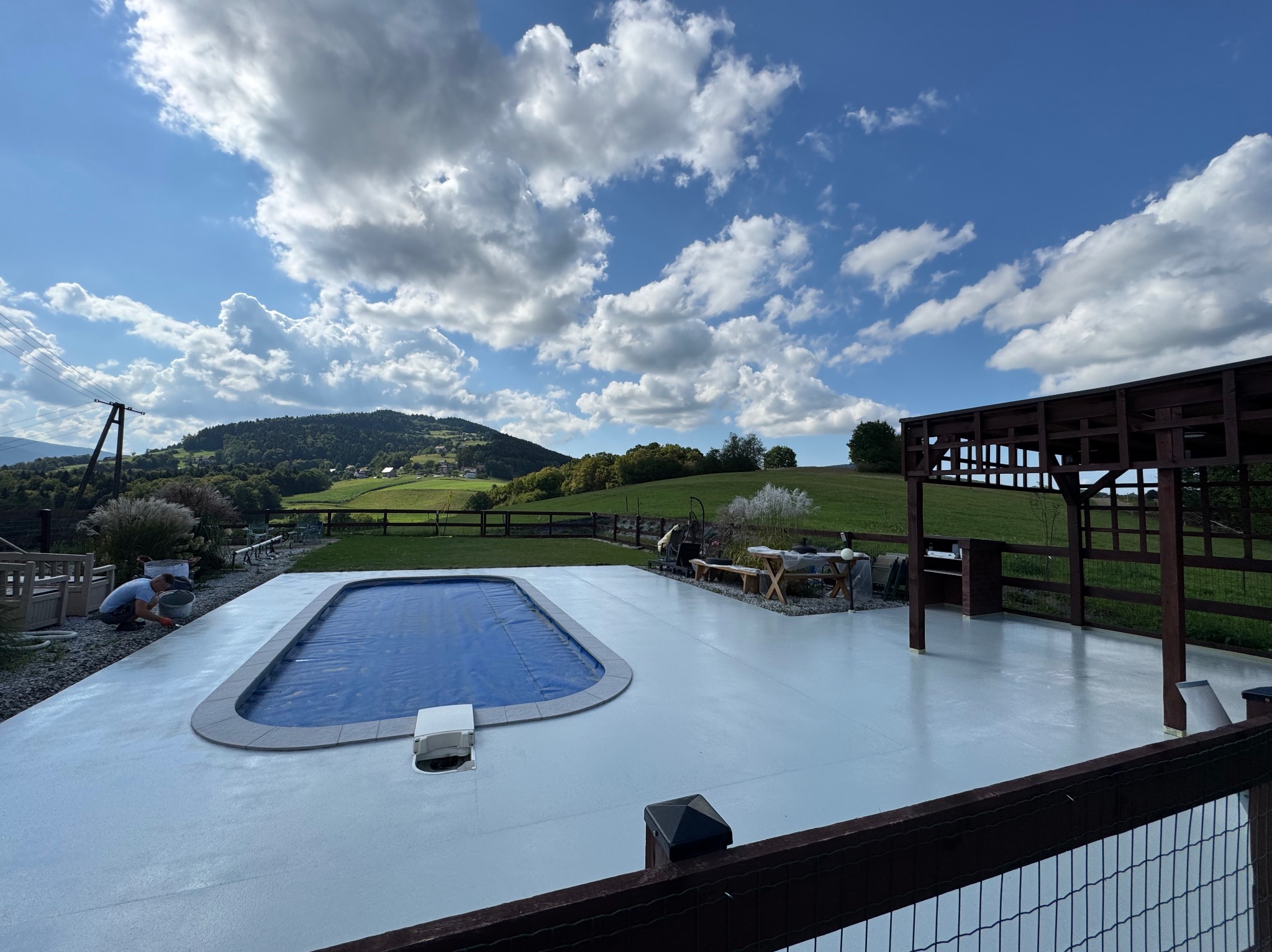 Jasnoszara posadzka żywiczna wokół basenu na zewnątrz, z widokiem na zielone wzgórza i błękitne niebo z chmurami. Osoba aplikuje materiał przy basenie.