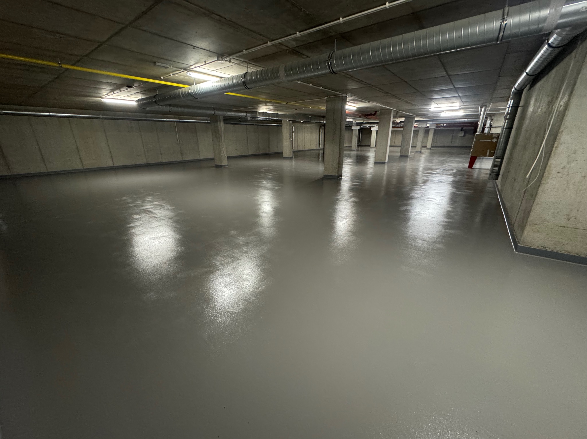 Gładka, szara posadzka żywiczna w przestronnym, surowym wnętrzu z betonowymi kolumnami i widoczną instalacją wentylacyjną pod sufitem. Minimalistyczny design.