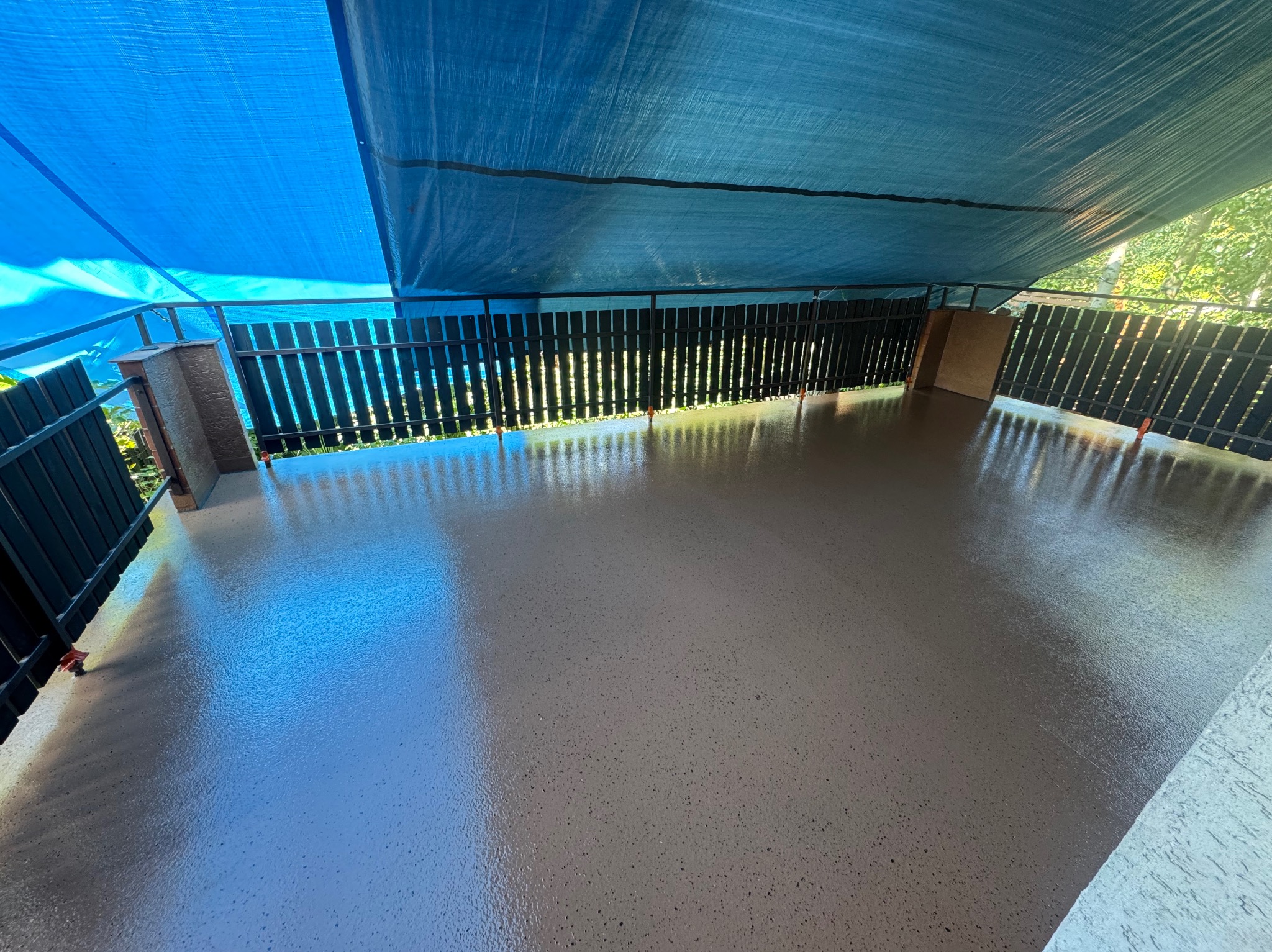 Błyszcząca, beżowa posadzka żywiczna na balkonie, zabezpieczona niebieską plandeką. Widoczne czarne barierki i fragment zieleni w tle. Świeżo wylana, gładka powierzchnia.