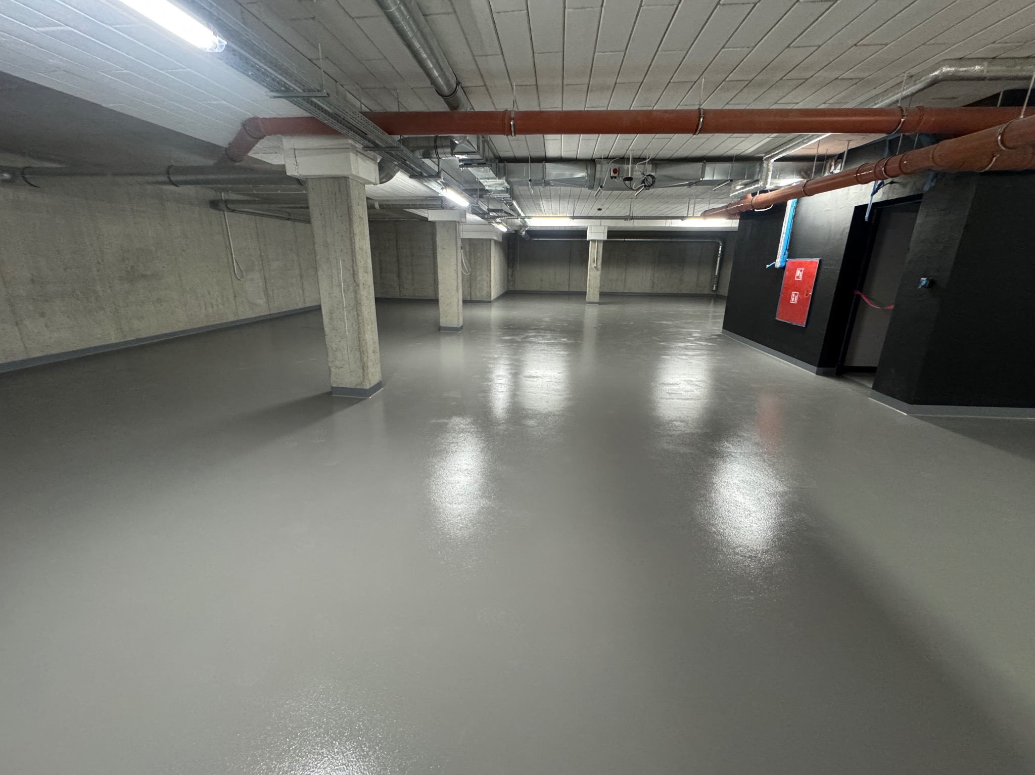Szara, gładka posadzka żywiczna w przestronnym, industrialnym wnętrzu z betonowymi ścianami i kolumnami. Widoczne rury pod sufitem i czarne elementy wykończeniowe.