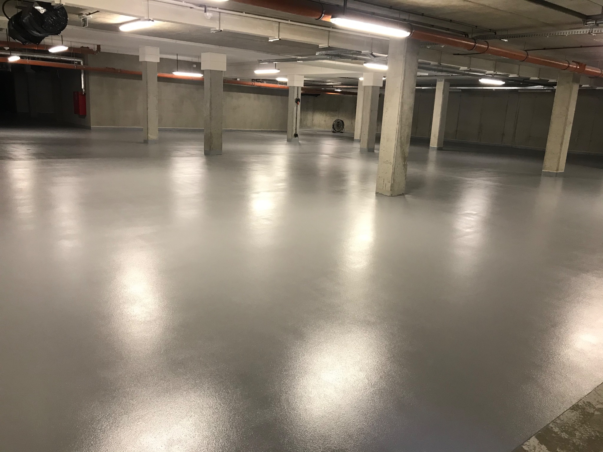 Szara, błyszcząca posadzka w garażu podziemnym, oświetlona lampami jarzeniowymi, z widocznymi betonowymi kolumnami i instalacjami pod sufitem.