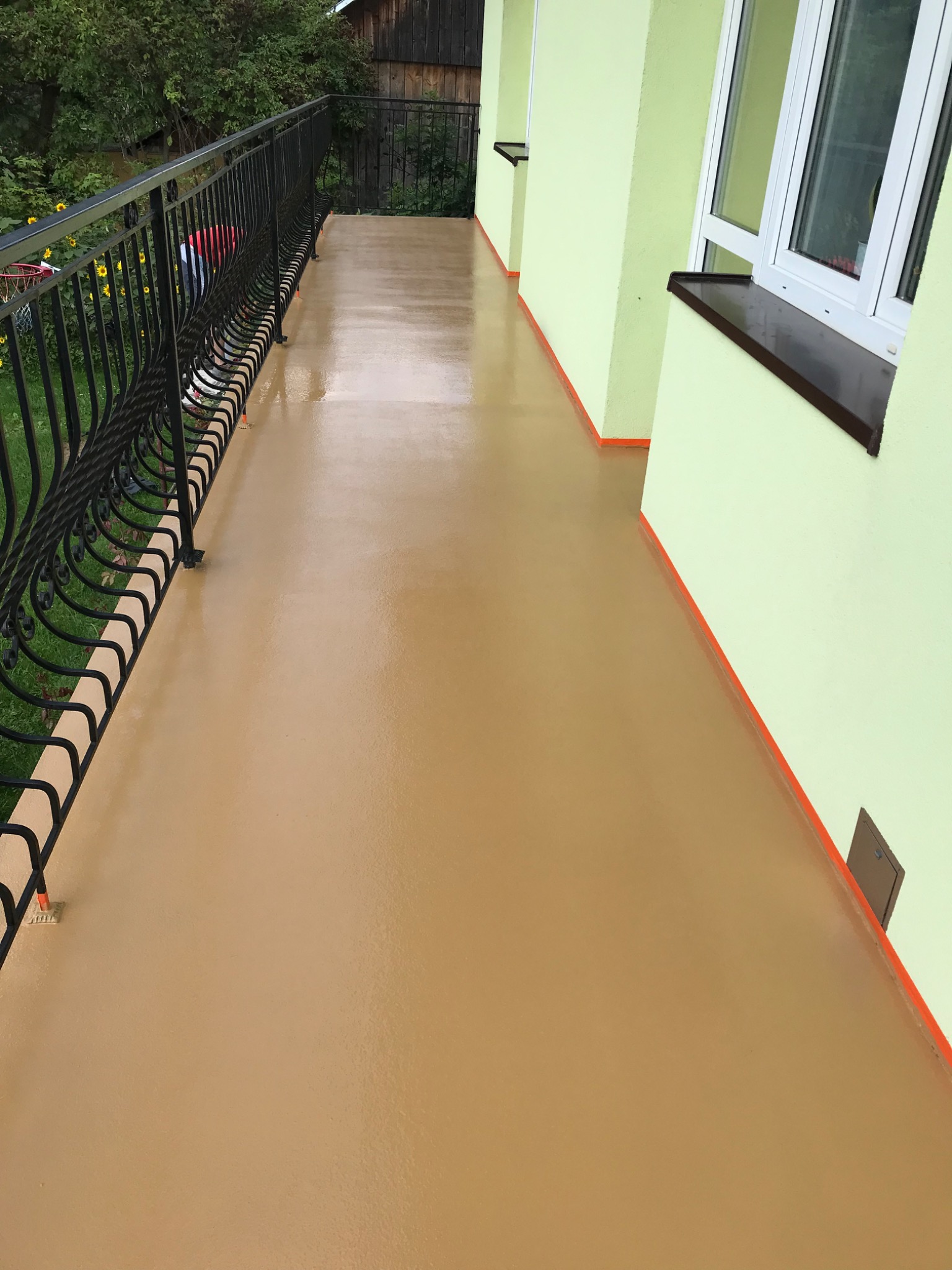 Balkon z beżową, jednolitą posadzką, zabezpieczoną czarną, kutą balustradą. Ściany budynku w kolorze pistacjowym z brązowymi detalami. Widok na zieleń ogrodu.