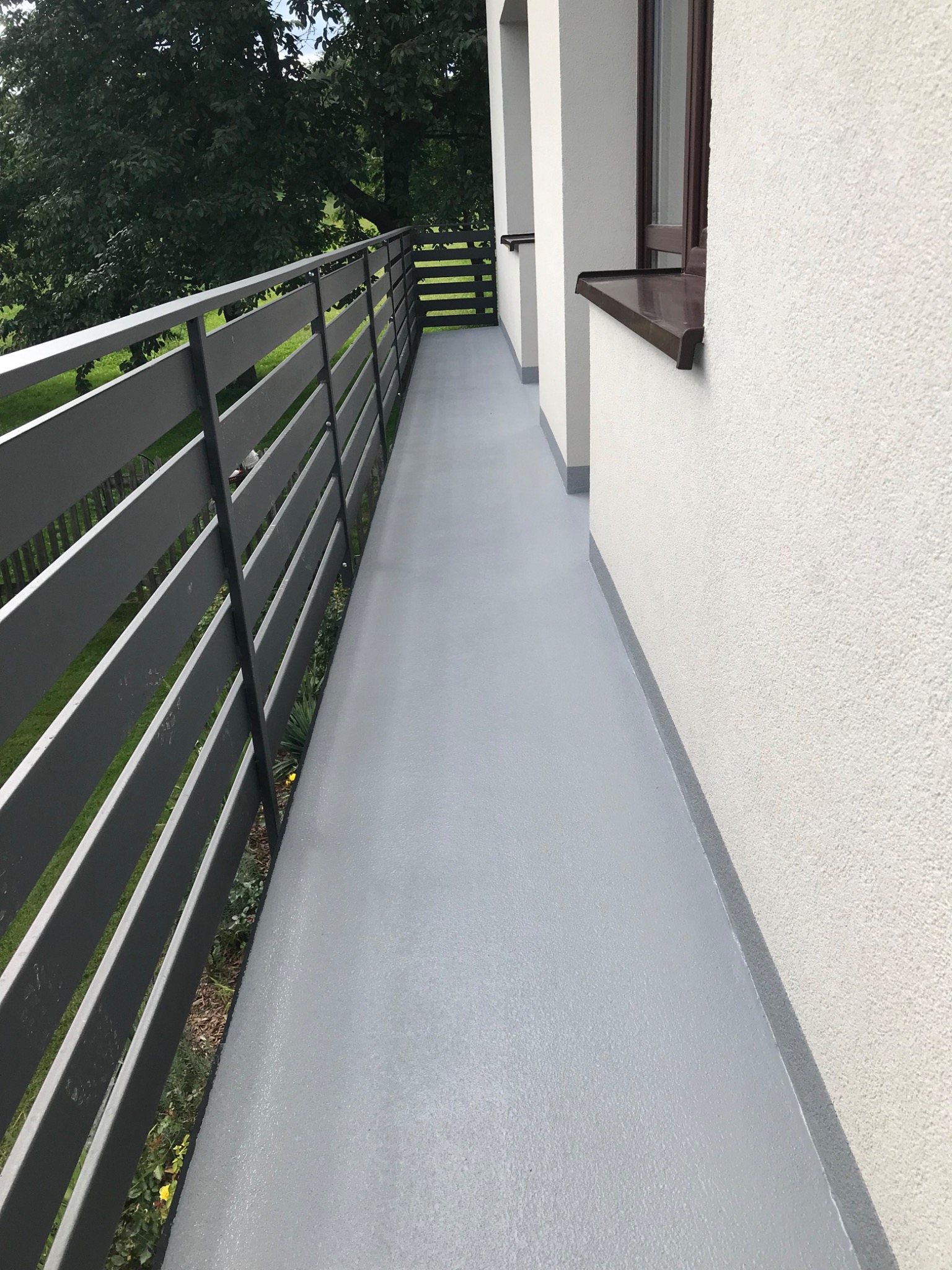 Szary balkon po renowacji z gładką powierzchnią, widoczne detale wykończenia przy ścianie i balustradzie, tło z zielenią drzew.