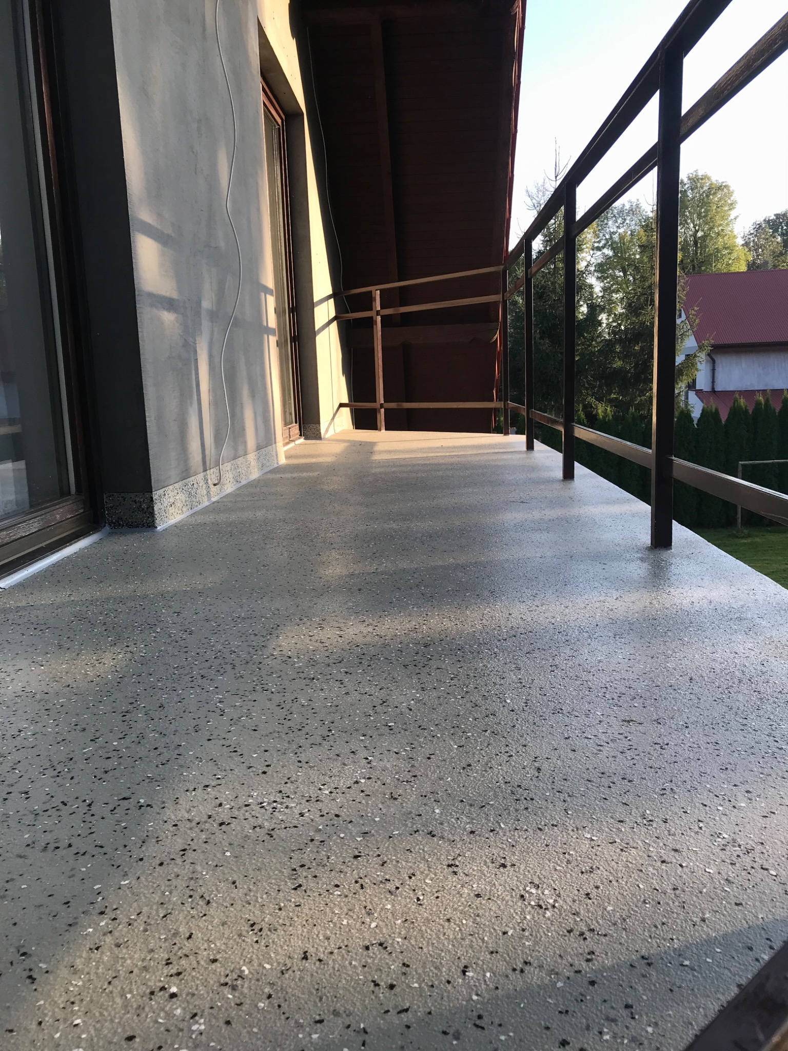 Szary balkon z żywiczną posadzką z widocznymi drobnymi, czarnymi i białymi kamyczkami, metalowa balustrada, widok na ogród w tle, promienie słońca padające na powierzchnię.