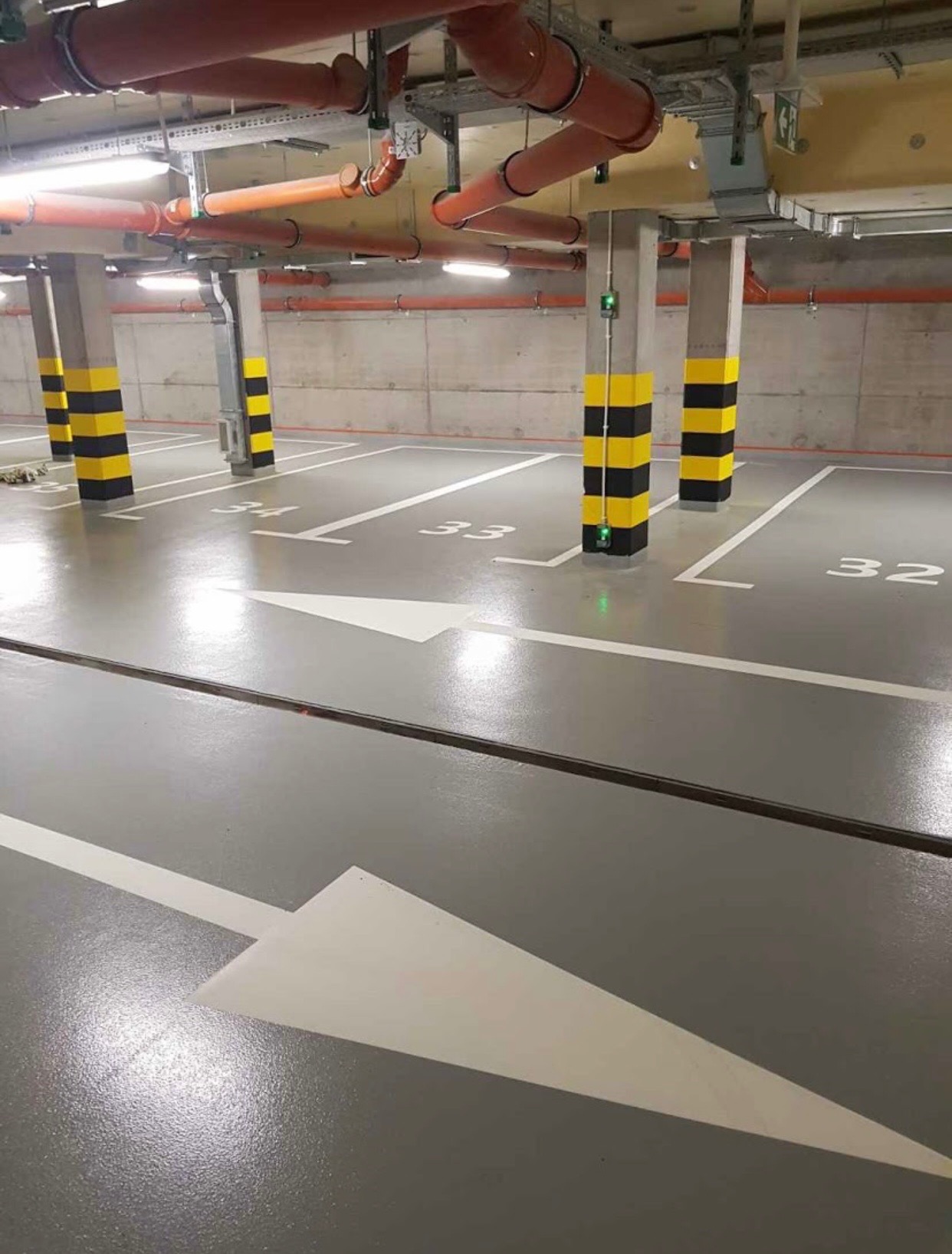 Szara posadzka w garażu podziemnym z numeracją miejsc parkingowych i strzałkami kierunkowymi, filary pomalowane w czarno-żółte pasy ostrzegawcze, instalacje wentylacyjne pod sufitem.