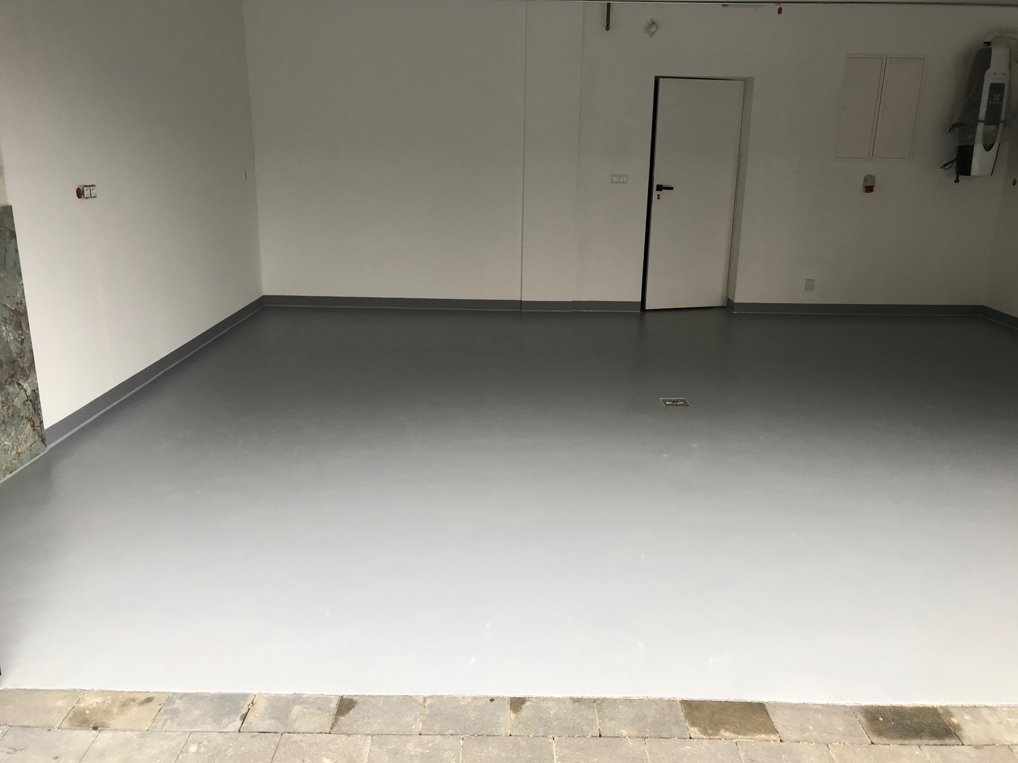 Jasnoszara, gładka posadzka w garażu z białymi ścianami i szarymi listwami przypodłogowymi. Widoczne drzwi wejściowe do garażu oraz brama garażowa z napędem.