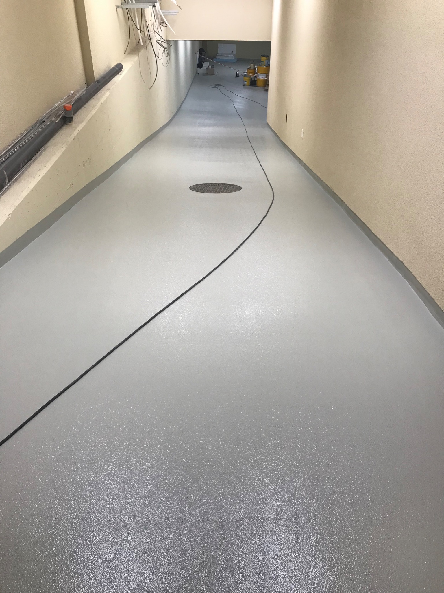 Długi korytarz z nową, gładką, szarą podłogą. Widoczne puszki z farbą w oddali i kabel na podłodze. Ściany beżowe z fakturą.