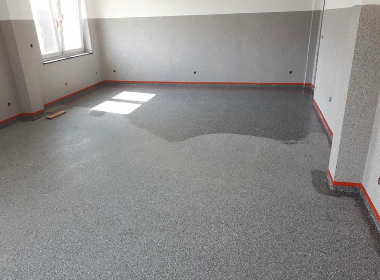 Szara, świeżo wylana posadzka żywiczna z widocznymi refleksami światła dziennego z okna, pomarańczowe listwy przypodłogowe, szare ściany z widocznymi otworami po kołkach.