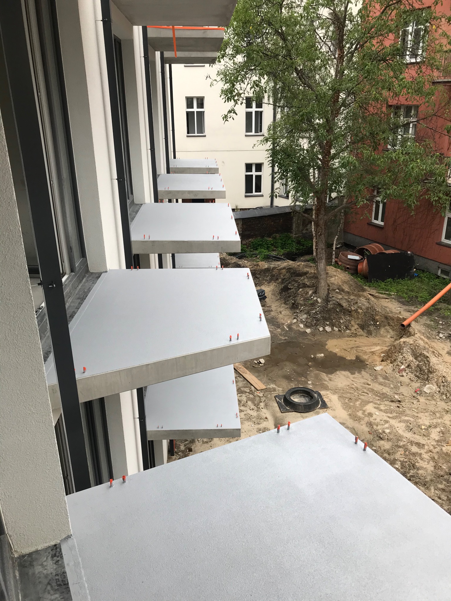 Nowe, szare balkony z betonu architektonicznego z pomarańczowymi elementami mocującymi wystającymi z powierzchni, widok z góry na plac budowy z drzewem i elementami kanalizacji w tle.