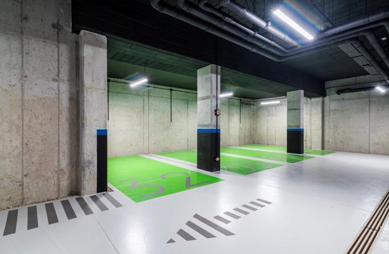 Wnętrze parkingu podziemnego z posadzką w jasnym odcieniu szarości, wyznaczonymi na zielono miejscami parkingowymi dla osób niepełnosprawnych, betonowymi ścianami i kolumnami oraz czarnym sufitem...