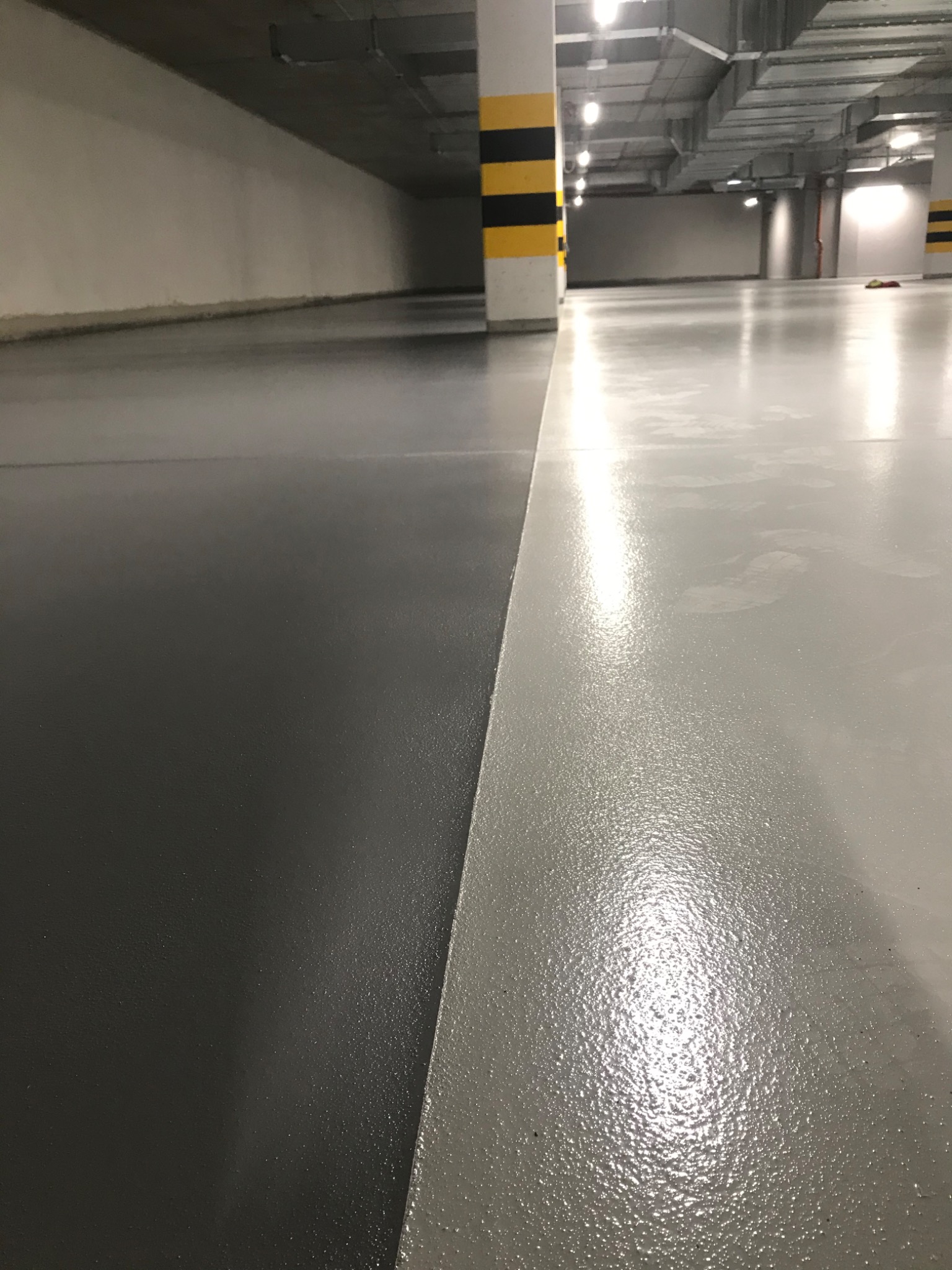 Dwie powierzchnie podłogowe w garażu podziemnym, jedna w kolorze szarym, druga czarna, oddzielone wyraźną linią; widoczny słup z żółto-czarnymi pasami.