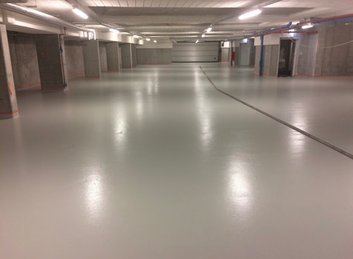 Jasnoszara, gładka posadzka w garażu podziemnym z betonowymi kolumnami. Widoczne rury pod sufitem i brama garażowa w oddali.