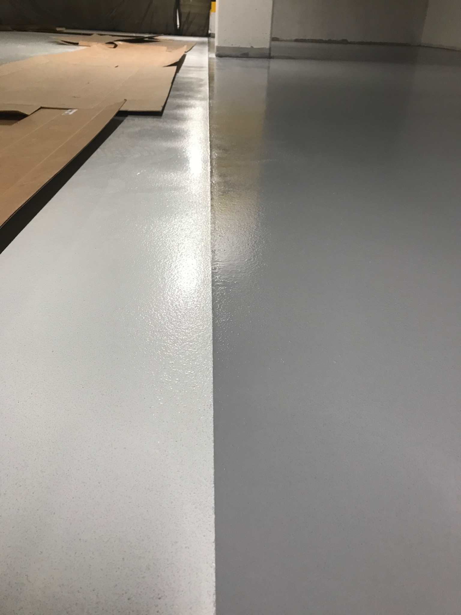 Świeżo wylana, błyszcząca posadzka żywiczna w dwóch odcieniach szarości, z kartonem ochronnym przy ścianie.
