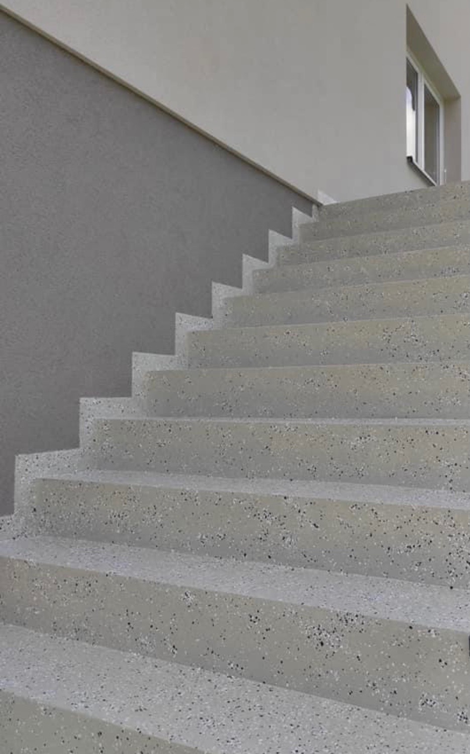 Schody zewnętrzne wykończone szarą, nakrapianą posadzką, prowadzące do budynku z szarą elewacją i oknem.