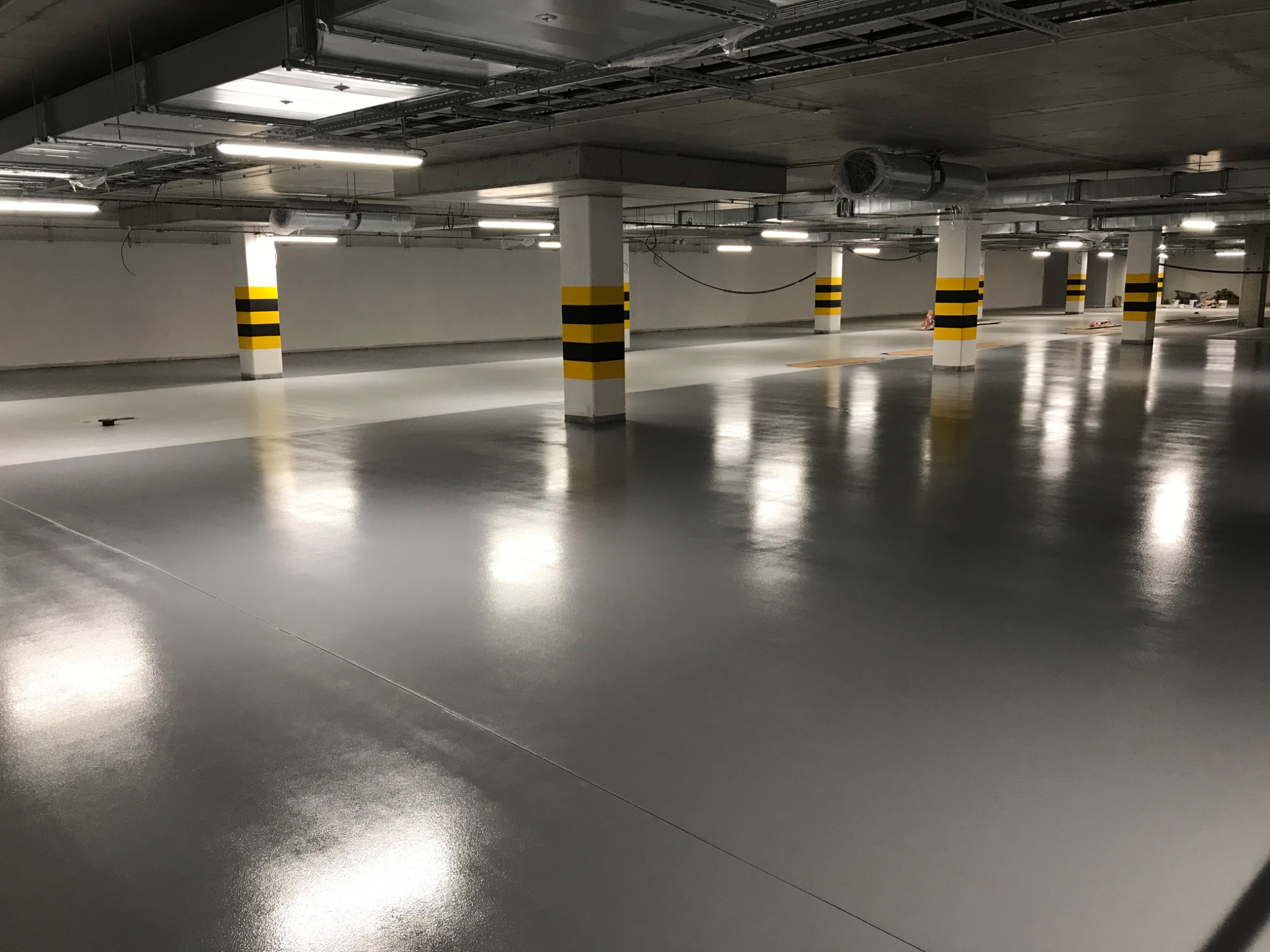 Szara, błyszcząca posadzka w garażu podziemnym z żółto-czarnymi pasami na filarach, widoczne oświetlenie sufitowe i instalacje.