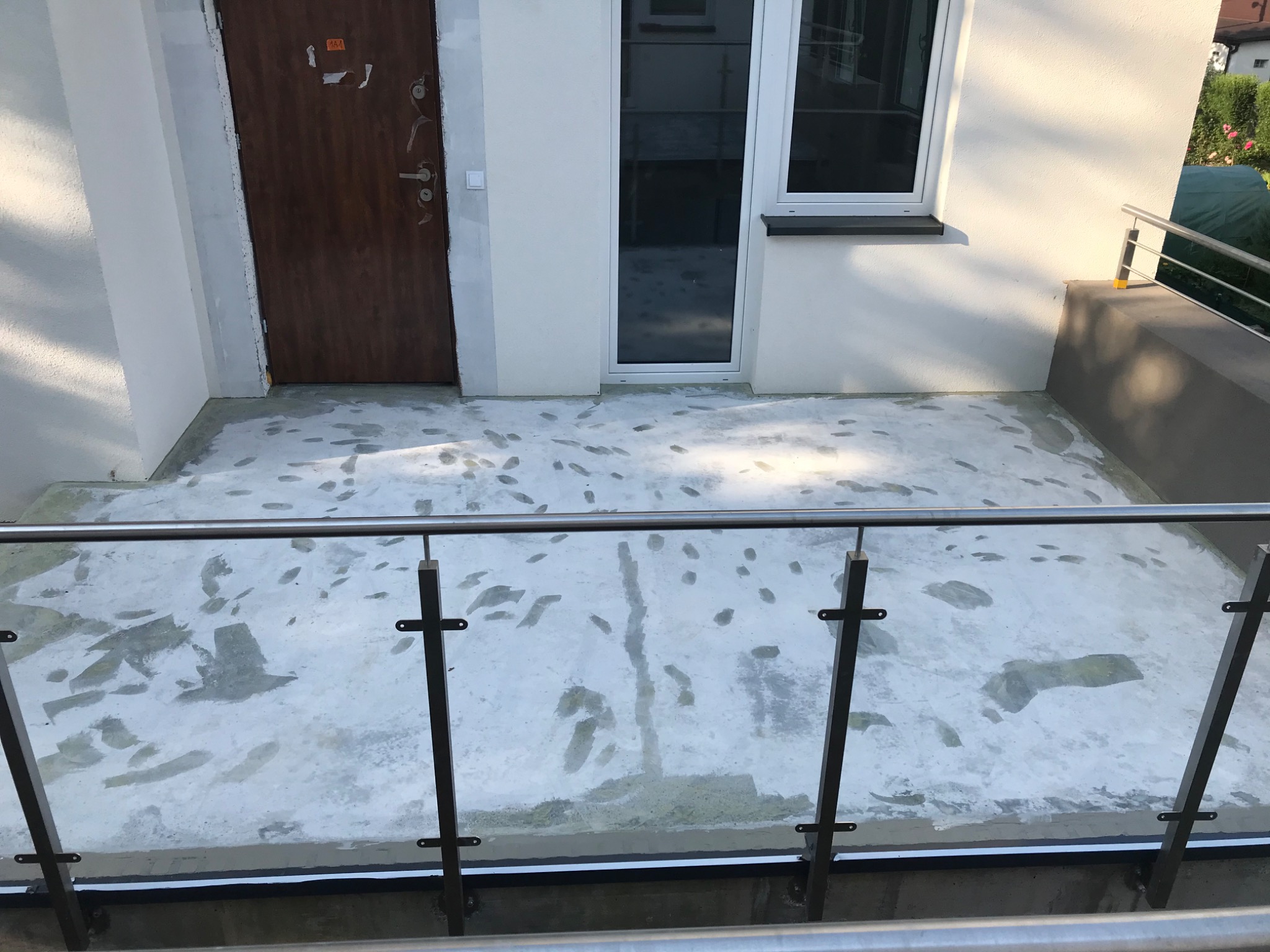 Biała posadzka na balkonie w trakcie prac, widoczne ślady stóp i niedociągnięcia, balustrada ze stali nierdzewnej z elementami szkła, drzwi wejściowe i okno w tle.