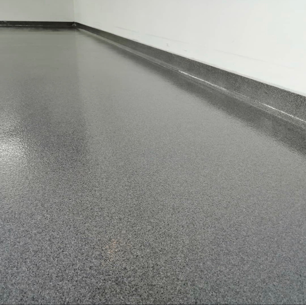 Szara, błyszcząca posadzka z drobnym ziarnem, widok z niskiej perspektywy w narożniku pomieszczenia, przy listwie przypodłogowej.