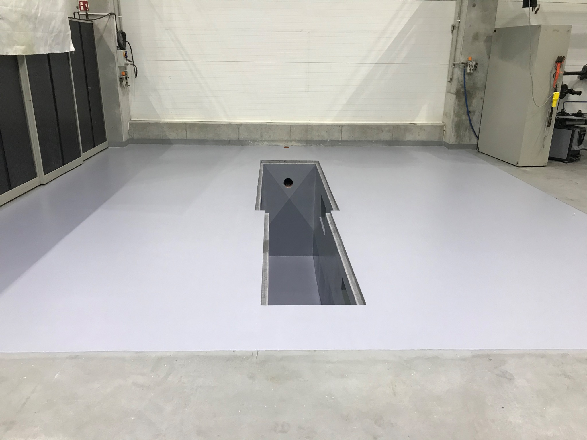 Jasnoszara, gładka posadzka w hali przemysłowej z kanałem rewizyjnym na środku. Widoczne instalacje na ścianach i szafy narzędziowe w tle.