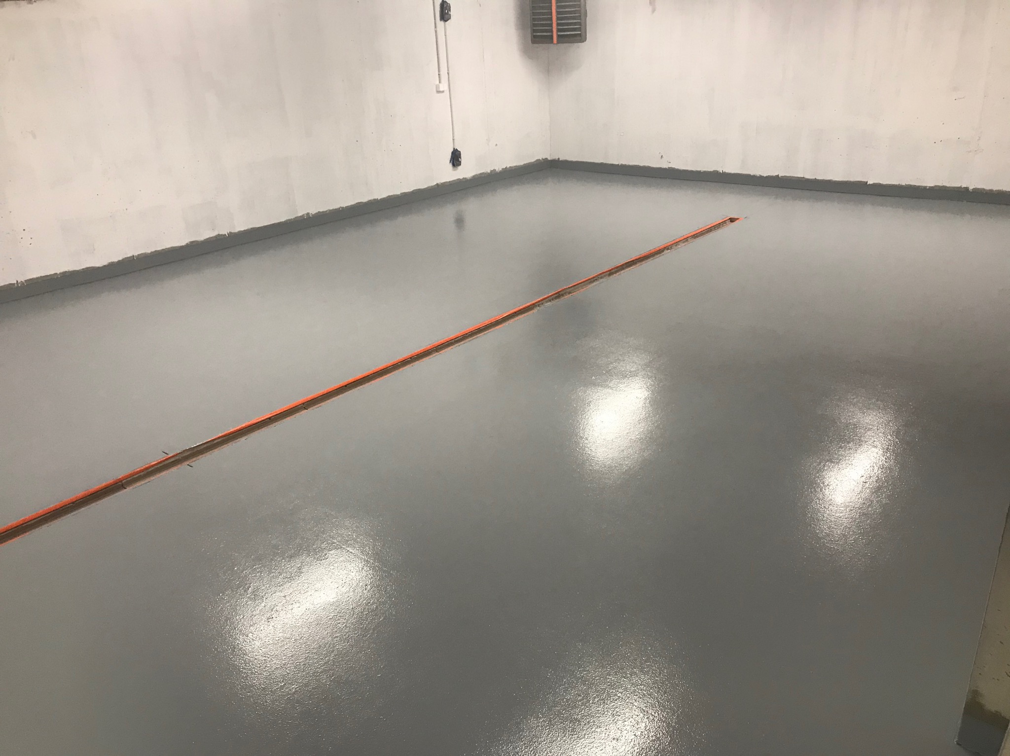 Świeżo wylana, szara, błyszcząca podłoga w pomieszczeniu; widoczna listwa dylatacyjna i fragment niepomalowanej ściany.