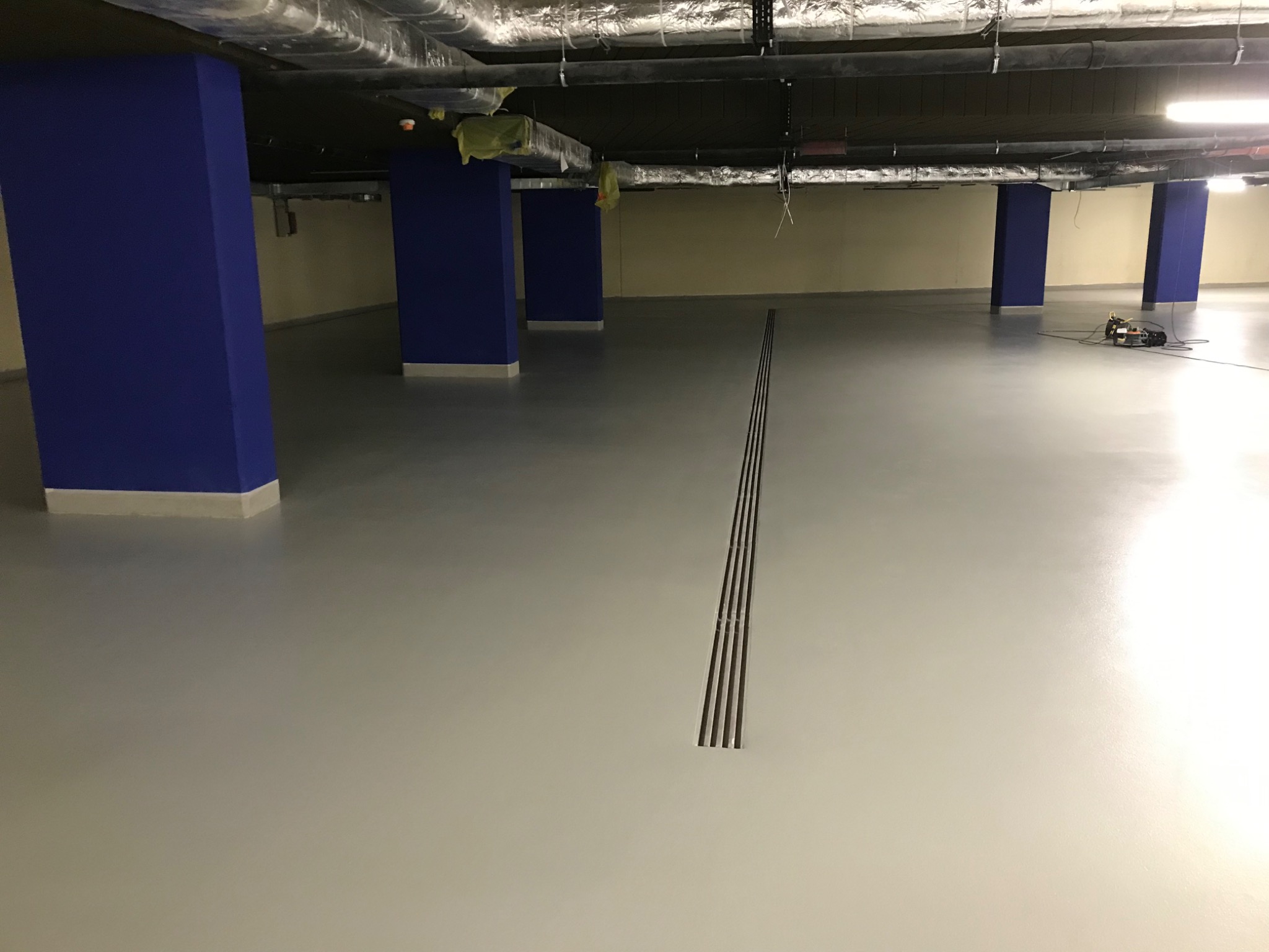 Jasnoszara, gładka podłoga w hali garażowej z niebieskimi filarami i kratką odpływową. Widoczne instalacje pod sufitem i sprzęt do polerowania podłogi.