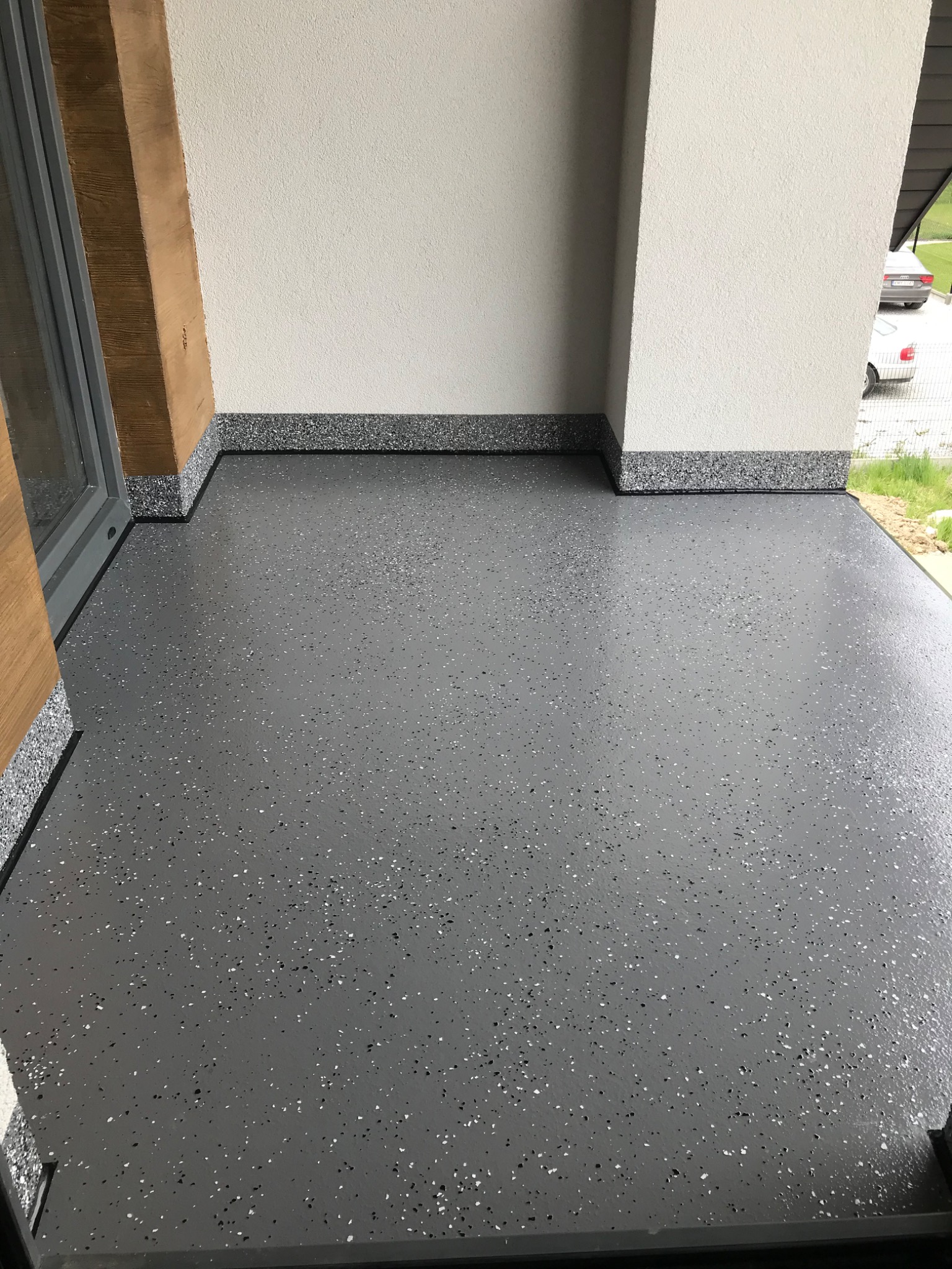 Szara, błyszcząca podłoga z drobnymi, czarno-białymi plamkami, wykończona listwą przypodłogową z tego samego materiału, widok z góry na fragment tarasu z elementami stolarki okiennej i ściany...