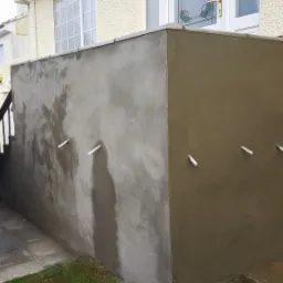 Surowa elewacja budynku po nałożeniu tynku, widoczne białe rurki wentylacyjne, fragment drewnianej szopy i schodów w tle.