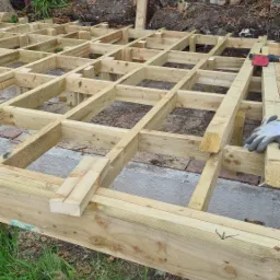 Konstrukcja nośna z impregnowanego drewna pod budowę tarasu, widoczny częściowo ułożony ruszt, narzędzia pracy i rękawica ochronna, w tle podbicie pod wyższą kondygnacją budynku.