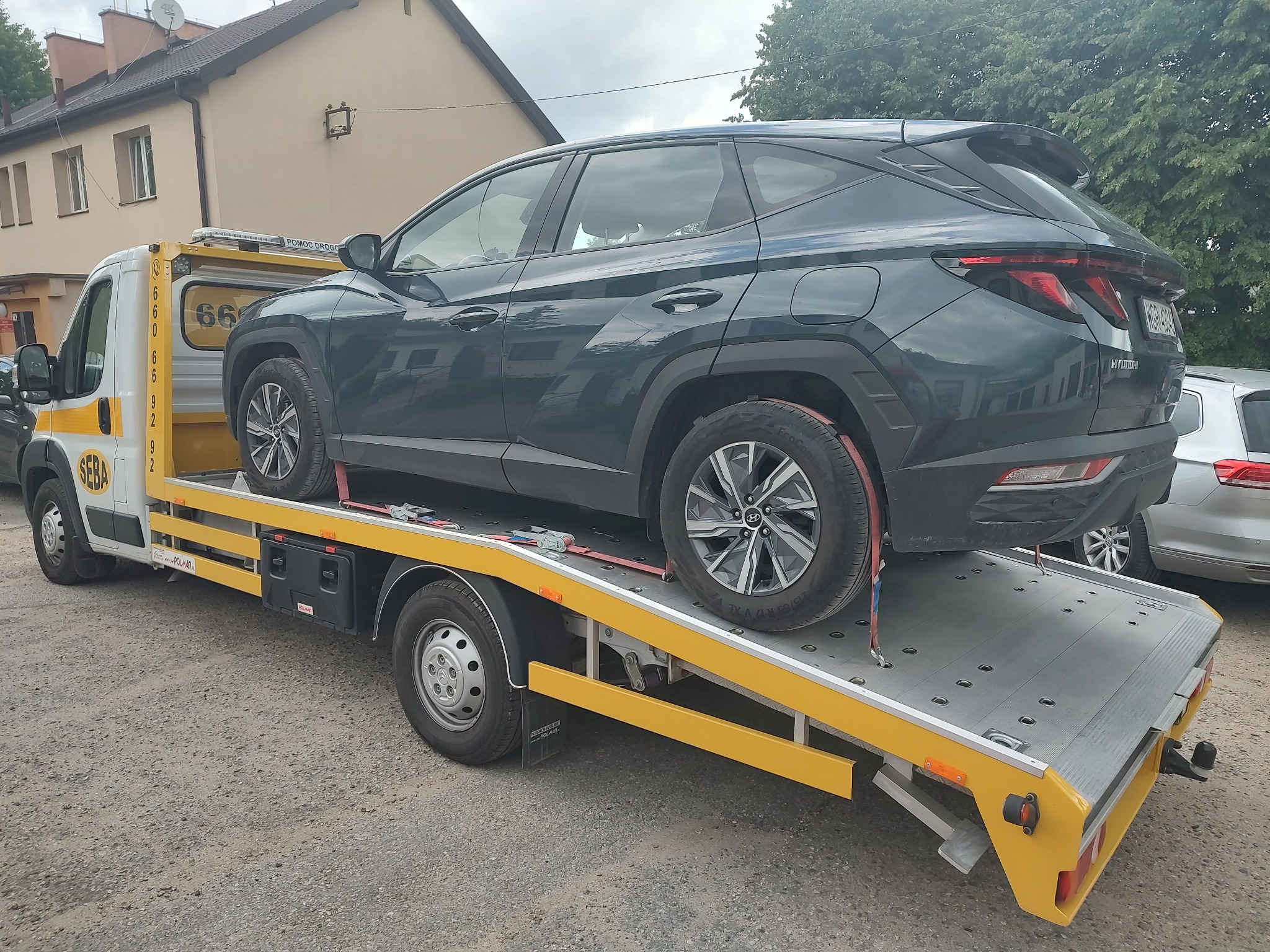 Szary samochód Hyundai Tucson zabezpieczony pasami transportowymi na żółtej platformie pojazdu pomocy drogowej z widocznym numerem 660669292, na tle budynku.